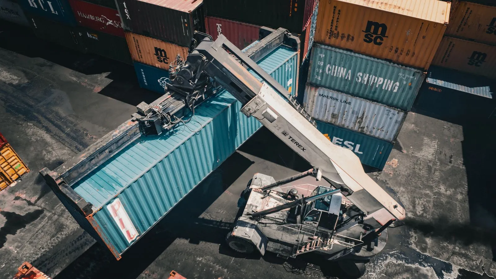 Aerial view of a reach stacker lifting a blue shipping container in a port yard, next to stacks of containers from different shipping lines