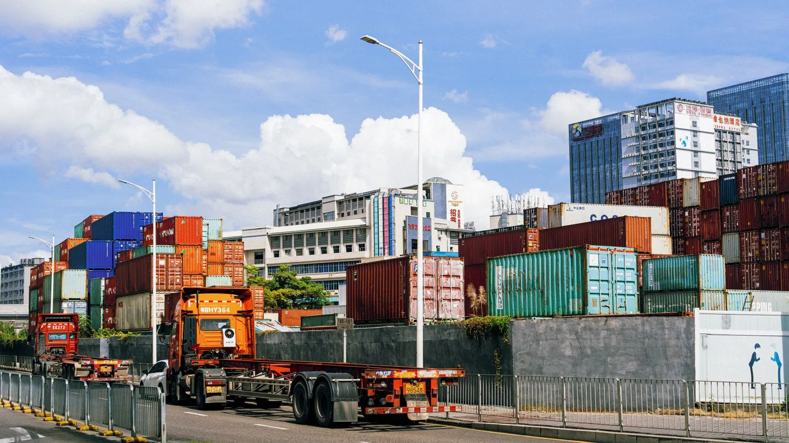 Stacks of shipping containers in a container yard, with two container trucks on the road in front