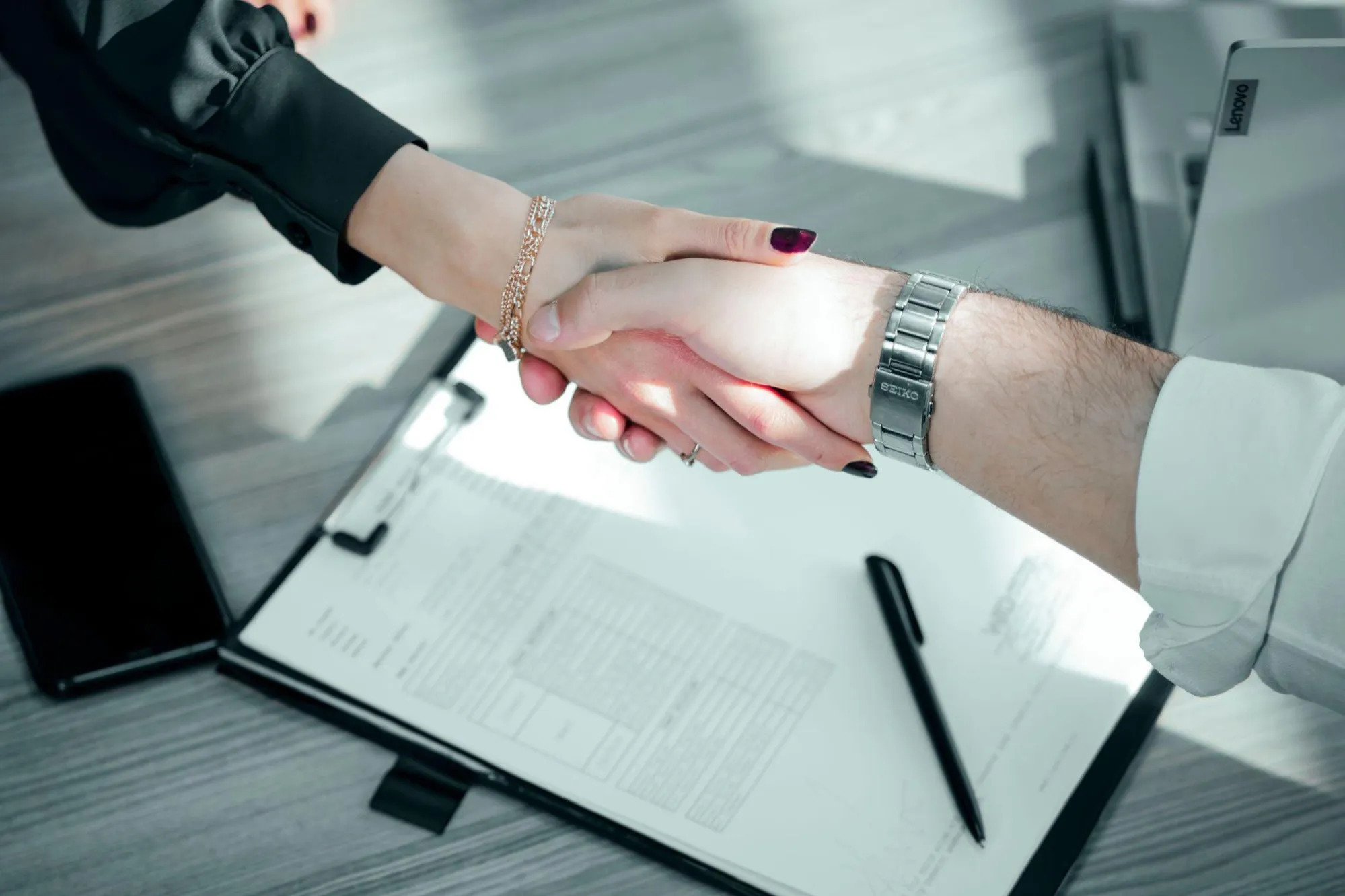 Two people shaking hands over a printed form on a clipboard, with a pen resting beside it