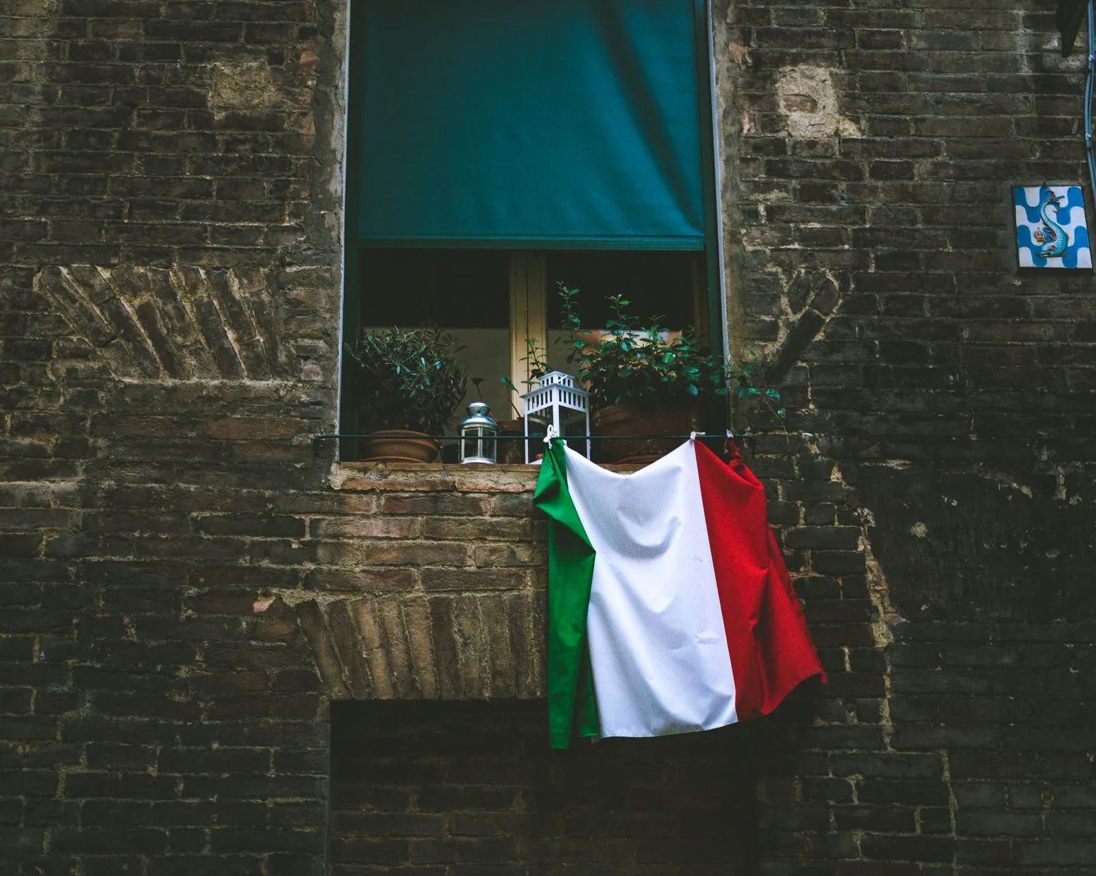 An Italian flag hanging from the sill of a window with potted plants and a lantern, set in an old brick wall