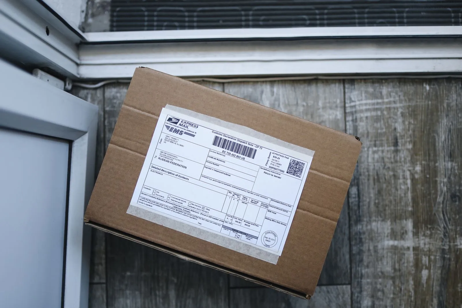 A cardboard parcel with a postal customs declaration label taped to its lid, propped against a weathered wooden wall