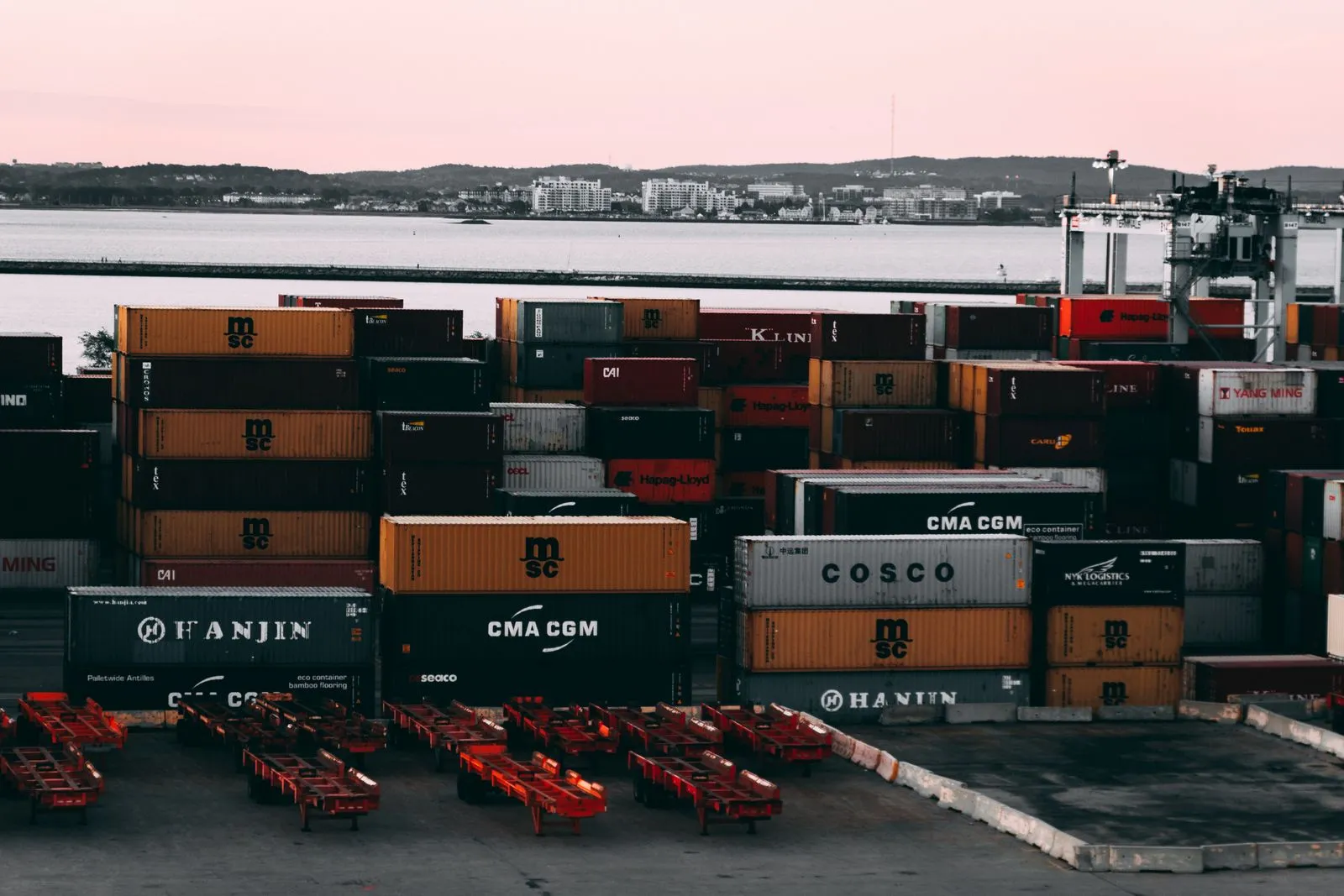 Stacked shipping containers in a port at dusk, with the water and a coastal town behind them