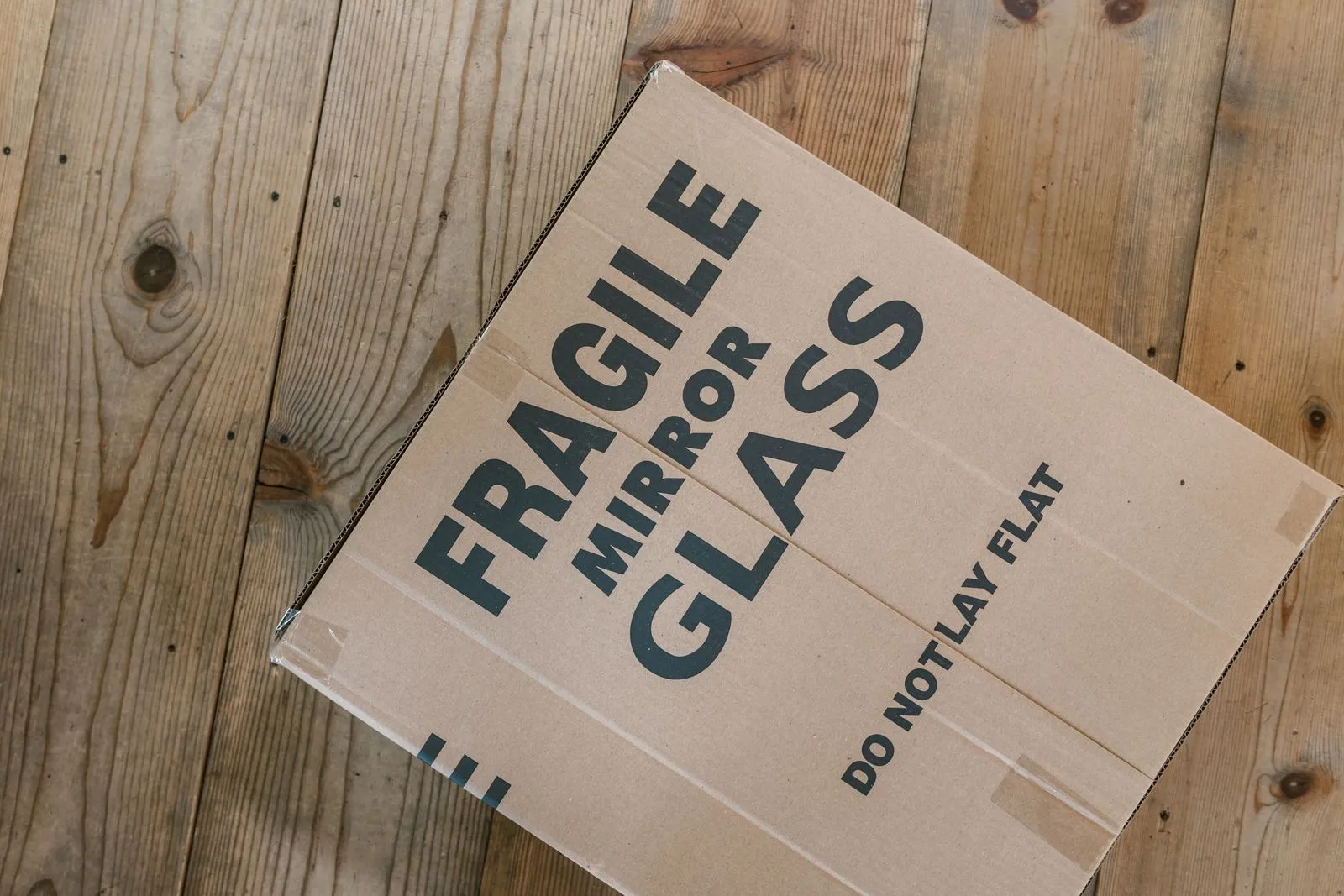 A flat cardboard box on a wooden floor, printed in large letters with the words FRAGILE, MIRROR, GLASS and DO NOT LAY FLAT