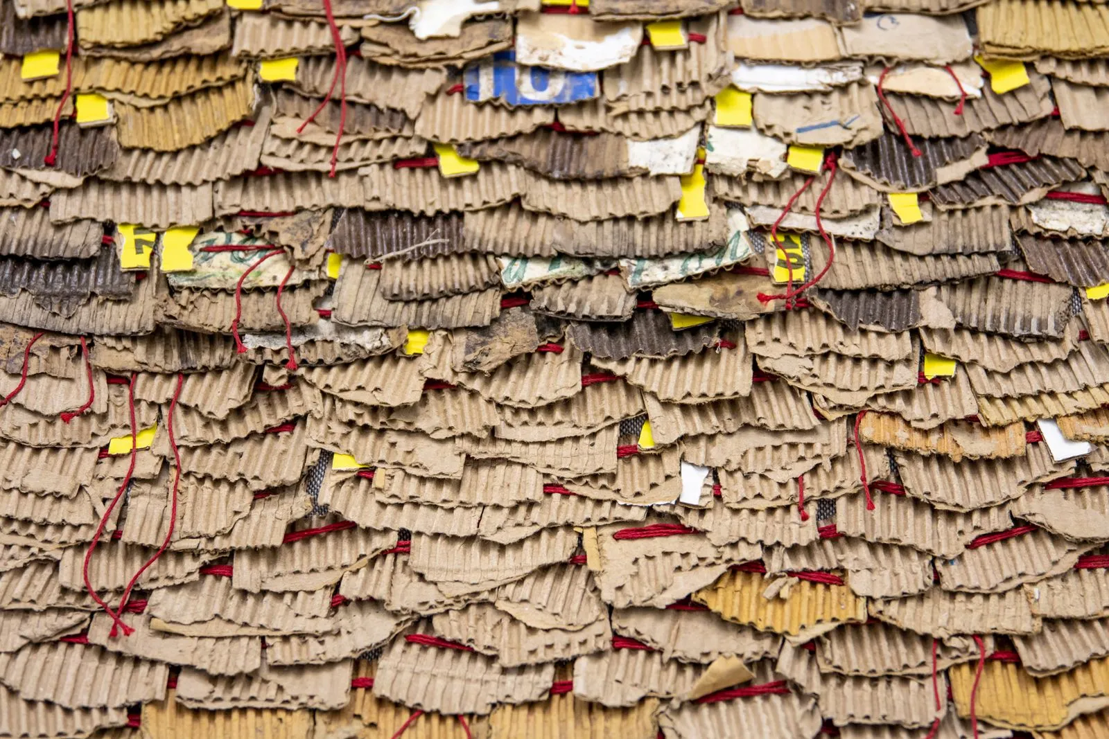 Layers of torn corrugated cardboard scraps stitched together with red thread, with fragments of coloured printing among them
