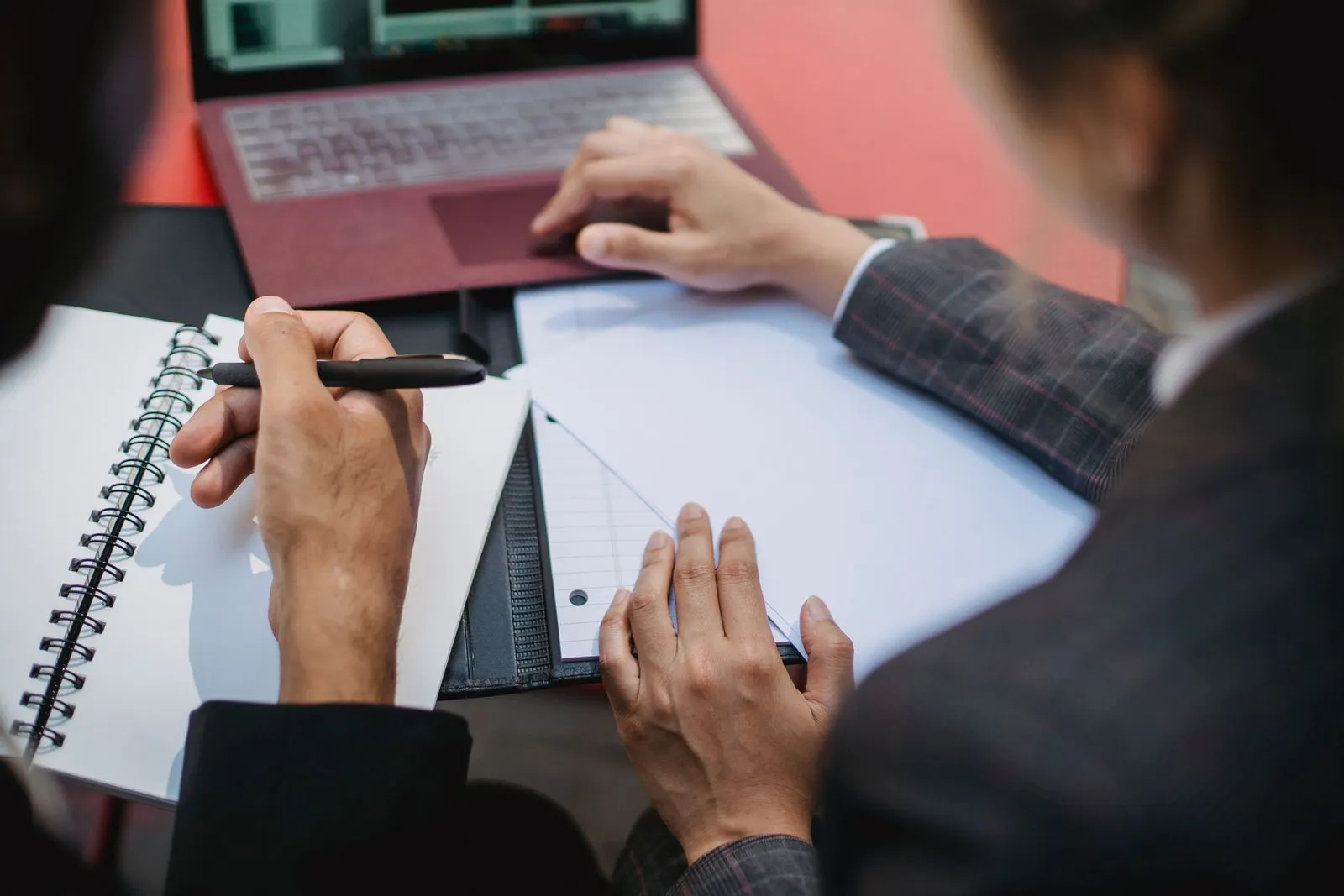 Two people at a desk going through printed documents and a notebook beside an open laptop