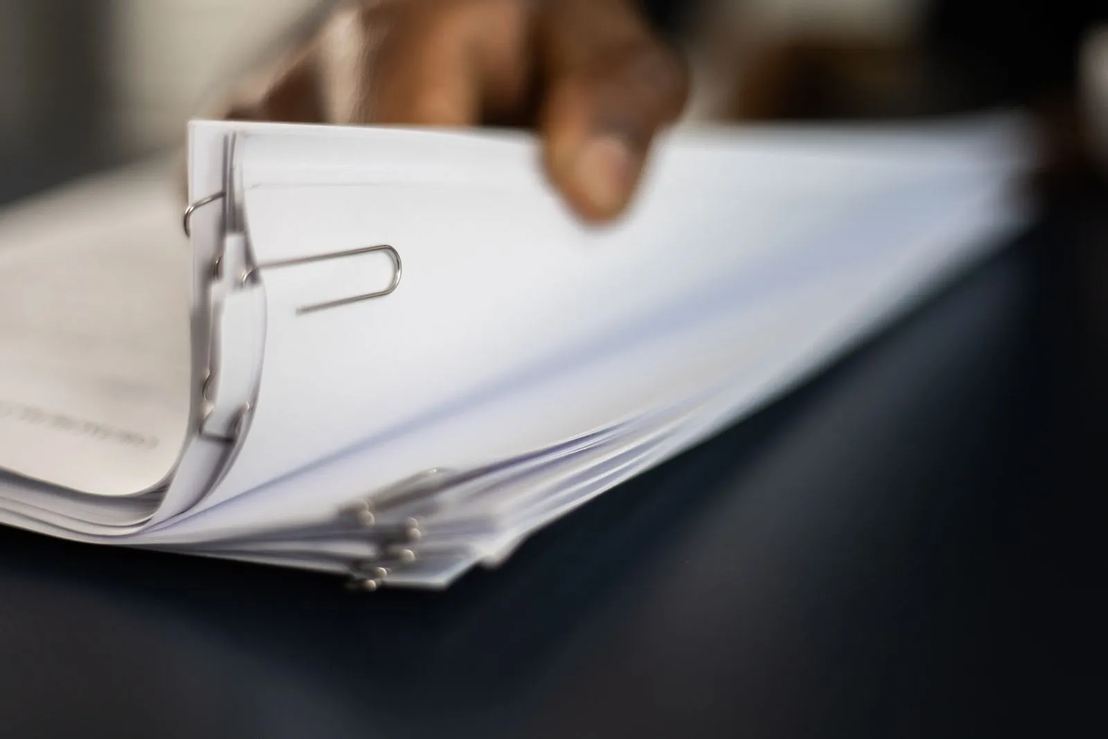 A hand leafing through a thick stack of white documents held together with paper clips