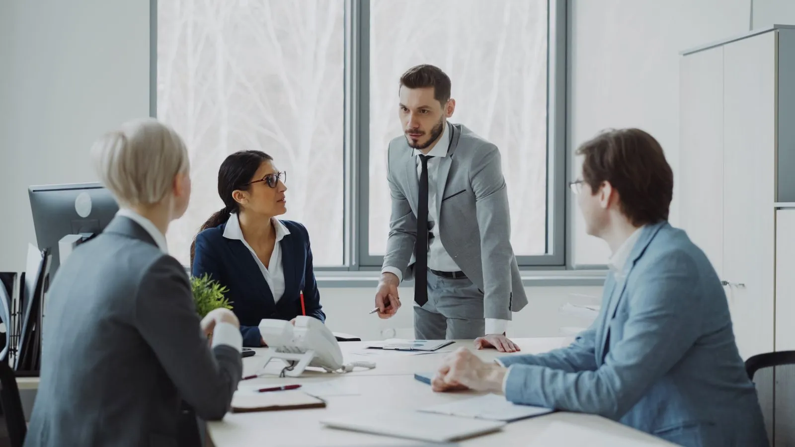 Four colleagues in business attire around a meeting table in a bright office, listening to a man standing and speaking