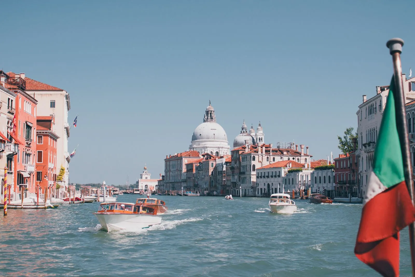 Motorboats on the Grand Canal in Venice heading towards the domes of Santa Maria della Salute, with an Italian flag on a pole in the foreground