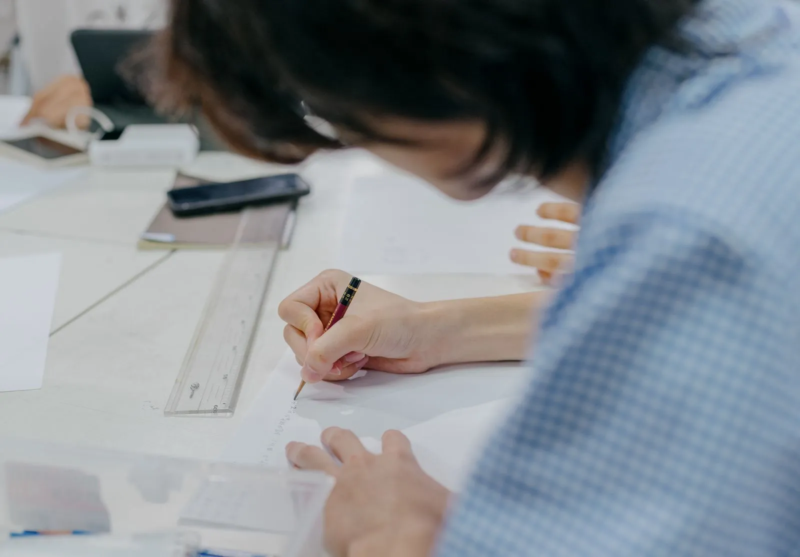A person in a checked shirt bent over a white desk, writing on a sheet of paper with a pencil next to a ruler
